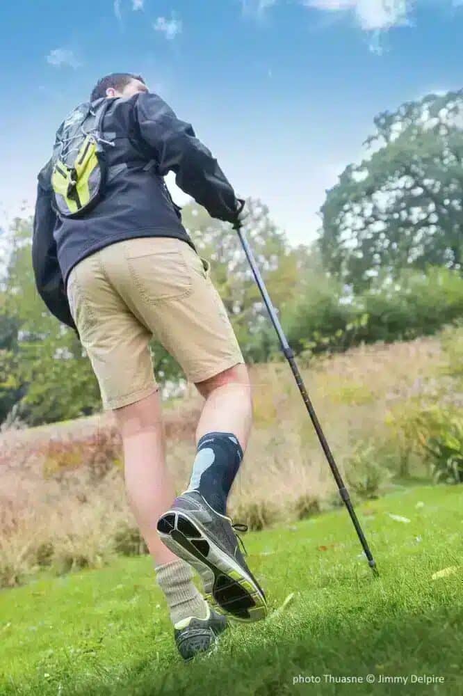 Ankelortos Silistab Achillo 25 Person med blått ankelstöd och vandringsstavar promenerar i gräsbeklädd naturmiljö