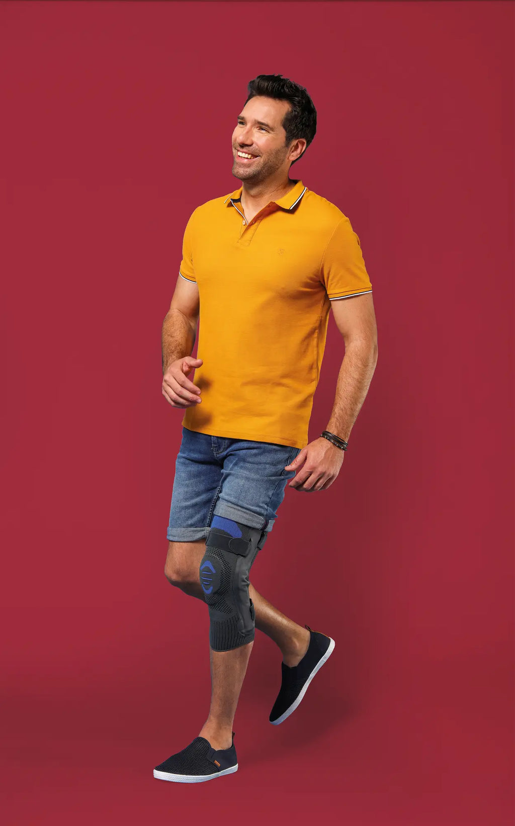 En man med knästöd Genu Dynastab går leende i en gul polo och jeansshorts.