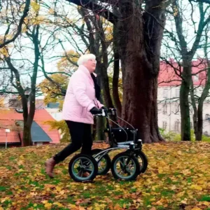 Person som går med rollator med stora hjul i en park på hösten bland fallna löv.