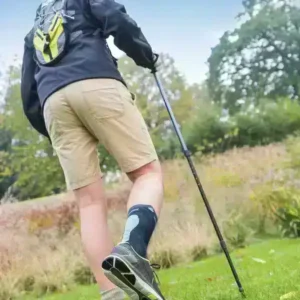 Person med blått ankelstöd och vandringsstavar promenerar i gräsbeklädd naturmiljö