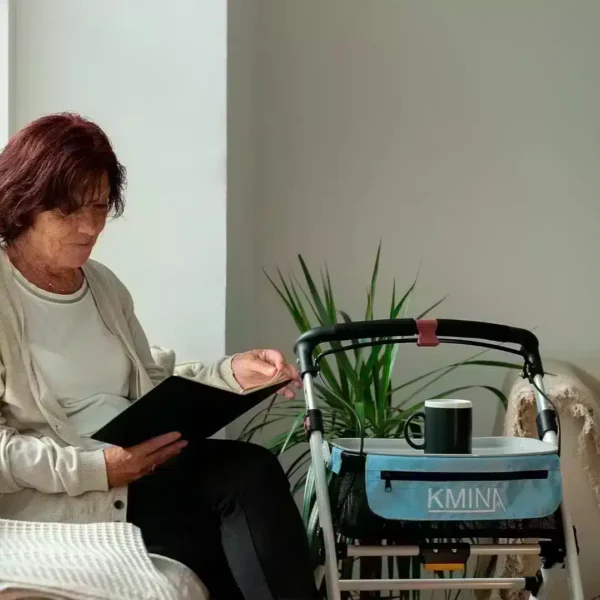 Kvinna som sitter i stol bredvid smal vit rollator med blå korg i hemmiljö.