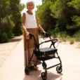 Person som promenerar med en rullator med fyra hjul på en solig gångväg omgiven av grönska.
